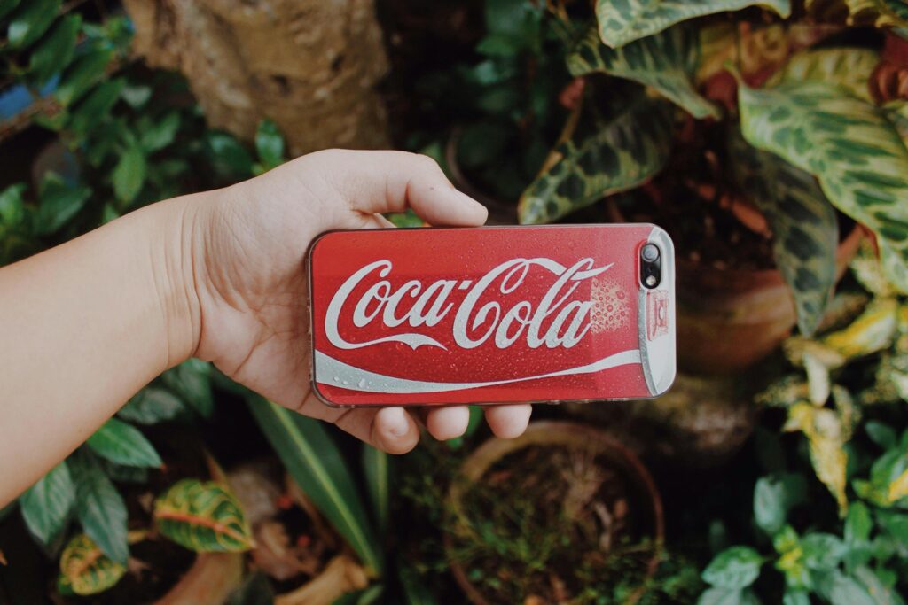 A hand holding a smartphone with a Coca-Cola case against a vibrant garden backdrop.